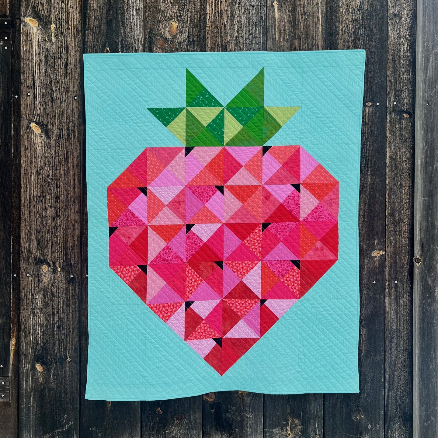 A quilt of a giant patchwork strawberry on a bright background. Quilt is hanging on the side of a wooden garage.